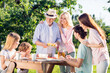 © deagreez - Multi-generation family enjoying an outdoor picnic with grandparents serving sandwiches while parents and children gather around a wooden table in a sunny summer garden