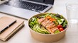 © thiago - A healthy, fresh salmon salad bowl with avocado and vegetables served for a quick office lunch next to a laptop, notebook, and a glass of water.