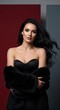 © GHM_STUDIO - Elegant woman with long wavy black hair wearing a black corset and faux fur coat against a red and grey studio background