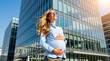 © miss irine - Pregnant woman wearing glasses stands in front of modern office buildings. She holds her belly, smiling upwards. Businesswoman enjoys her career and future.
