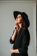 © Raisa - Stylish woman in black hat and blazer poses in studio. She thinks upward with hand on chin. Elegant look for fashion campaigns or beauty editorials.