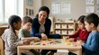© Alva - A smiling female teacher engages with four diverse young children, two boys and two girls, at a wooden table in a brightly lit classroom, playing with colorful educational blocks.