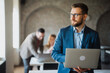 © Novak - Confident businessman holding a laptop with colleagues in the background.