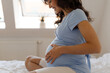 © Home-stock - Happy pregnant woman sitting on bed gently touching her baby bump and smiling while enjoying peaceful moments of pregnancy at home, side view