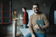© Novak - Confident businessman holding laptop with colleagues working in background