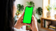 © MD Rashedul Islam - Hand holding mobile smartphone showing green screen chroma key mockup, dark hair woman in white shirt near indoor monstera plant and wooden bookcase for app ui ux design display