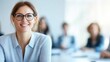 © Photography09 - Image A close-up portrait of a smiling woman wearing glasses and a light blue shirt. Background Blurred (bokeh effect) office environment with other people, suggesting a professional
