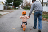 Orange bike wheels roll gently down quiet street. Child wears helmet, rides with trust and delight. Adult walks beside, hand steady, offering safety. Soft autumn leaves scatter beside pavement