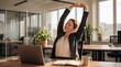 © Antonio - Businesswoman taking a break to stretch her arms above her head while sitting at her desk in a modern office, with a laptop and coffee cup nearby.