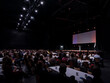 © Victor - A crowd of attendees fills a big convention hall with a bright stage screen glowing in the dark.