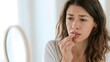 © suldev - A lifestyle portrait of a young woman in a bright, clean room, checking her mouth with her fingers while looking in a hand mirror, showing concern about the condition of her lips and teeth.