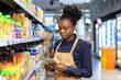 © Liubomir - Woman employee wearing an apron and working in a supermarket aisle, checking product stock and managing inventory with a modern digital tablet