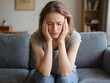 © Moopingz - A distressed woman sits on a couch, holding her head with both hands, expressing deep discomfort.