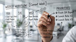 © Brokkk - Close-up of a hand writing a weekly workflow and priorities on a transparent glass board in an office setting.