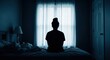 © Ariestia - Silhouette of a lonely woman sitting on a bed in a dark room facing a window, concept of depression and mental health