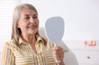 © New Africa - Senior woman looking in mirror at her teeth with dental implant at clinic