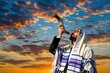 © Vadym - Jewish man with beard wears tallit prayer shawl blowing shofar ram horn. Dramatic sunset sky with orange clouds and blue sky background. Outdoors religious ceremony call.