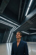 © f_bossa - Man wearing dark navy blazer looks upward within an expansive brutalist concrete hall featuring angular ceiling lines and long fluorescent tubes casting bright linear streaks across the floor.