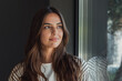 © Daniel - Thoughtful young brunette woman gazes out a window, bathed in soft natural light with a subtle reflection on the glass, conveying calm, introspection and quiet confidence in a casual setting.