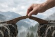 © Stocker - Man's hand carefully placing last wooden block to complete small bridge over deep crevice symbolizing shared effort problem-solving and trust with foggy peaks.