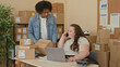 © Krakenimages.com - Woman on phone at desk with laptop and parcels while coworker reaches for boxes on a shelf in a building; small business teamwork busy.