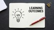 © Mudassir - Open notebook with learning outcomes text and a lightbulb icon placed on dark textured background