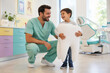 © SerPak - A dentist in green scrubs smiles while kneeling next to a young boy holding a large tooth prop. Friendly dental environment for children.