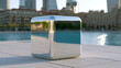© Tetyana - Shiny chrome cube sitting on a light tiled surface, prominently reflecting high rise buildings, historical structures, and the blue water of a major city