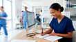 © Xenia - Smiling female medical assistant writing in notebook at desk. Professional nurse in scrubs working in busy hospital clinic with stethoscope and computer. Healthcare administration concept