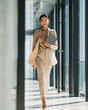 © syaifuddin - Confident successful Asian businesswoman walking through a modern glass office carrying a laptop, elegant beige business suit, warm natural sunlight, luxury corporate headquarters