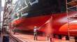 © Вадим Перегудов - Industrial workers painting the hull of a large ship in a dry dock. Men in protective gear using spray guns to apply red paint to a maritime vessel. Shipyard maintenance and repair
