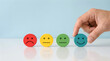 © MOMOTAJ - Hand selects happy green smiley face from row of colored emoticon discs showing customer satisfaction survey feedback in positive mood assessment experience.
