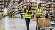 © A5 - Female warehouse worker pointing and holding a tablet while male colleague pushes a manual trolley with a box through a large distribution center with high metal shelving units.