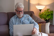 © luciano - Senior man with gray hair and beard comfortably sitting on a sofa, smiling while using modern laptop technology at home, embracing digital connection and online leisure