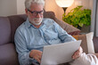 © luciano - Senior man relaxing on a comfortable sofa at home. Wearing eyeglasses while happily browsing the internet for leisure and engaging with online content using his laptop