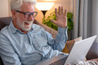 © luciano - Smiling senior man connecting with family or friends through a video call on his laptop. Actively waving a welcoming hand while sitting comfortably on a couch