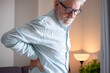 © luciano - Senior man with white hair and eyeglasses experiencing severe lower back pain. Holding his hand to his painful lumbar region while bending forward
