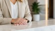 © olga_demina - A confident and poised professional woman in a modern office setting, sitting at a white marble counter with her hands clasped together, wearing a beige blazer and a white shirt, with a blurred green