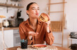 © Prostock-studio - Fast food at home. Excited young woman enjoying a flavorful burger, having junk food meal at home, sitting in kitchen interior, free space
