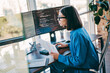 © deagreez - Female programmer at a sunny loft office coding on multiple monitors while reviewing documents