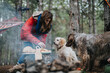 © qunica.com - A woman prepares food over a small campfire while two dogs wait nearby. The outdoor camping scene shows casual cooking and companionship in a wooded area.