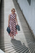© qunica.com - Woman wearing a pink polka dot dress and sunglasses walks up a sunlit textured ramp carrying a bag. The fashionable retro outfit and strong shadow create a chic urban street scene.