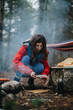 © qunica.com - Woman crouched at a campsite preparing food near a smoky campfire. A camper arranging bread and supplies on a rustic forest cooking area.