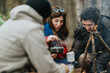 © qunica.com - A group of friends camping in the woods share hot tea around a small campfire. They laugh and pass enamel mugs while the kettle steams over the fire.