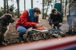© qunica.com - A woman and her friends camp in a mountain forest while she slices sausages on a cutting board. The group gathers near a small campfire beside a tent with a dog nearby.