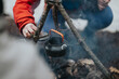 © qunica.com - A person lifts the lid of a small kettle suspended over a smoky campfire. The outdoor scene shows camping gear and a hot drink being prepared in a forest.