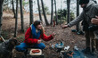 © qunica.com - Friends cook sausages over a campfire in a mountain forest while sharing smiles and warm clothing. A small group enjoys outdoor cooking and camping together with a dog nearby.