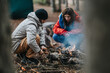 © qunica.com - Two friends crouch by a small campfire in a mountain forest on a cool day. The pair prepare wood and tend the fire while camping outdoors.