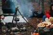 © qunica.com - A kettle hangs over a small campfire as people prepare bread and fruit at a forest campsite. The scene shows outdoor cooking and casual camping food prep with smoke rising from the fire.
