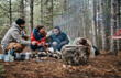 © qunica.com - Three friends sit around a small campfire roasting food and smiling with their dog nearby. A tent and camping gear are visible as they relax together in the pine woods.